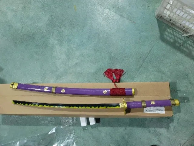 Katana Enma morada de Zoro de One Piece decorativa con saya morada, sageo rojo y hoja con detalles encima de caja. Vendida por Espadas y más